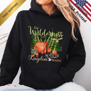 Może przedstawiać: Czarna bluza z kapturem z grafiką o tematyce kempingowej. Projekt przedstawia pomarańczowy namiot, ognisko, drzewa oraz słowa "Wilderness Tested Kingdom Bound" w zielonych i złotych literach. Bluza jest drukowana w USA.