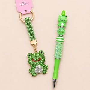 May include: A green frog-themed keychain and pen set. The keychain has a green strap with a gold ring and a rhinestone-covered frog charm. The pen is green with a frog topper and rhinestone accents. The keychain tag reads "mint".