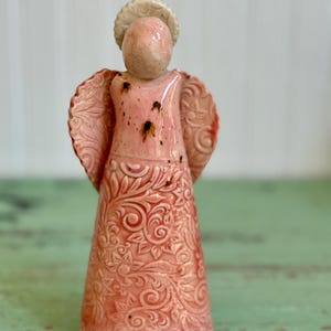 May include: A ceramic angel figurine in shades of pink and cream. The angel has detailed wings and a halo. The body of the angel is adorned with a floral pattern. The figurine is a decorative item.