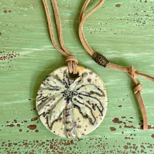 May include: A ceramic pendant necklace featuring a palm tree design. The round pendant is cream-colored with a brown palm tree illustration. The necklace has a tan leather cord and a decorative metal bead.