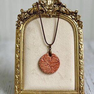 May include: A round, speckled peach-colored ceramic pendant featuring a debossed floral pattern. The pendant hangs from a thin, dark brown cord and is secured by a bronze-toned metal bail, creating a simple and distinct piece of jewelry.