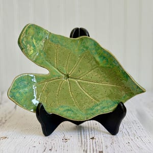 May include: A ceramic dish shaped like a large leaf with a glossy finish. The surface features a mottled light and dark green glaze with visible dark speckles and embossed veins radiating from the center. The dish is held by a black stand.