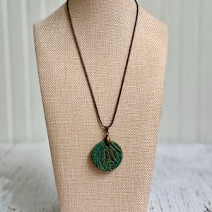 May include: A circular pendant featuring a textured green surface with dark leaf-like impressions, suspended from a thin dark brown cord. The pendant is attached to the cord by a small bronze-toned metal bail, creating a simple necklace design.