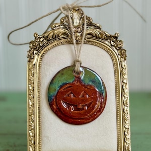 May include: A circular ceramic ornament featuring a carved jack-o'-lantern face with a glossy orange and green glaze. The ornament hangs from a natural twine string centred within an ornate, rectangular gold-toned metal frame with floral details.