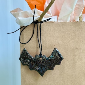 May include: A dark, glazed ceramic bat ornament with intricate embossed patterns, suspended by a thin black string from the handle of a brown paper gift bag containing orange and white tissue paper. The bat features a glossy, multi-toned finish.
