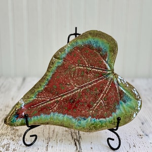 May include: A heart-shaped ceramic dish molded in the form of a leaf. The center features a deep red glaze with green speckles, while the edges are finished in a vibrant green and blue gradient. The dish rests on a black metal display stand.