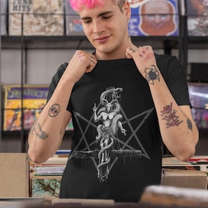 May include: A black t-shirt featuring a graphic print of a goat-like figure with snakes wrapped around it. The figure is on a cross with a pentagram behind it. The t-shirt is being worn by a person with pink hair.