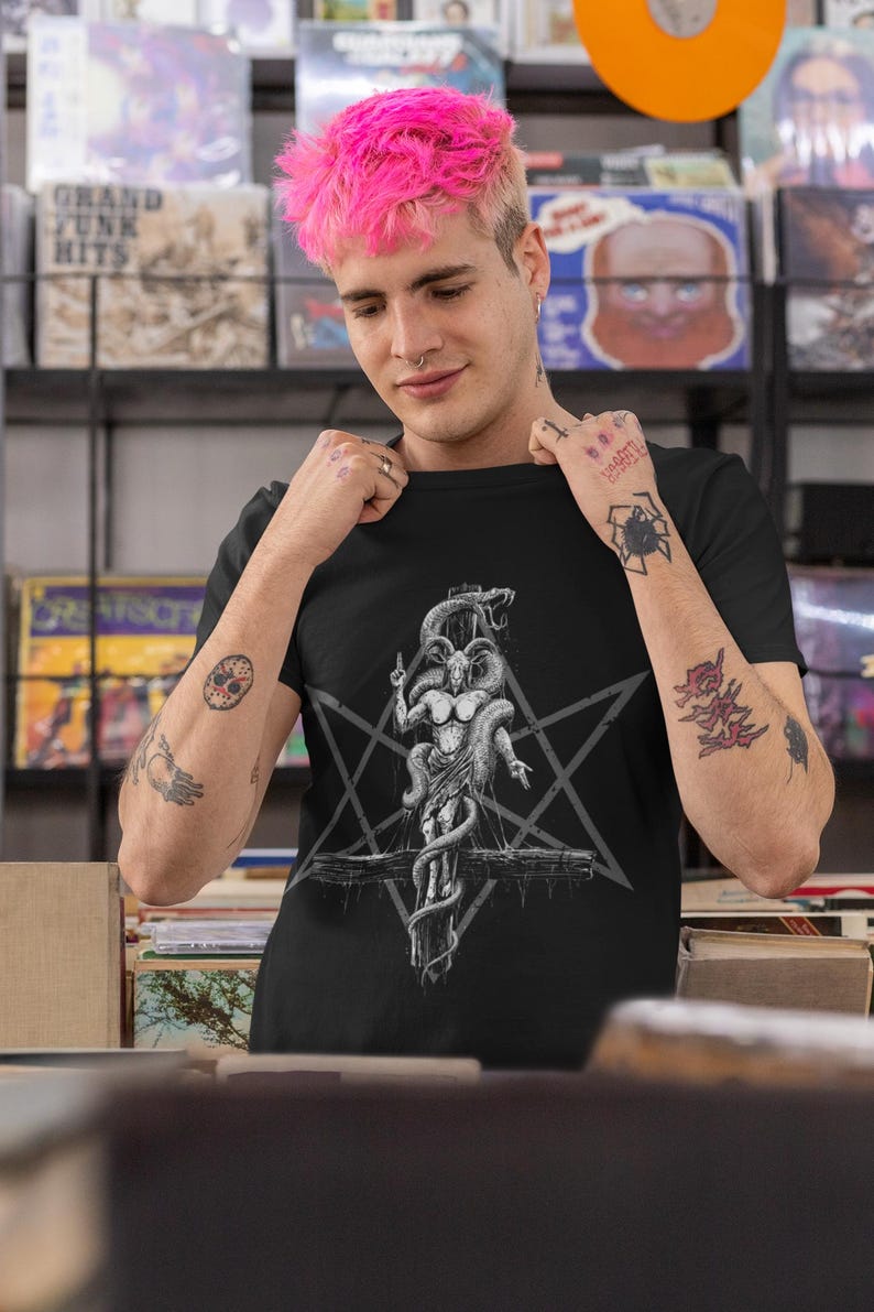 May include: A black t-shirt featuring a graphic print of a goat-like figure with snakes wrapped around it. The figure is on a cross with a pentagram behind it. The t-shirt is being worn by a person with pink hair.