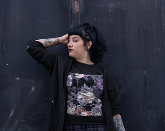 May include: A black t-shirt with a floral print featuring a skull and a dark background. The shirt is worn by a person with black hair and a black jacket. The person is standing in front of a dark metal door.
