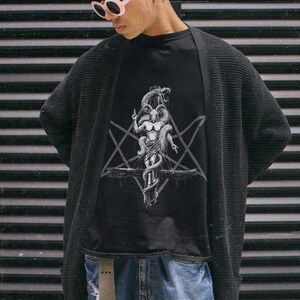 May include: A person wearing a black t-shirt with a white graphic of a goat-like figure with horns and a snake wrapped around it. The figure is standing on a cross with a pentagram behind it. The person is also wearing a black cardigan and blue jeans.