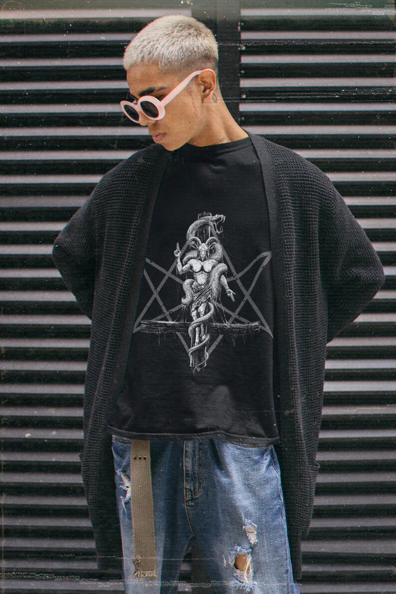 May include: A person wearing a black t-shirt with a white graphic of a goat-like figure with horns and a snake wrapped around it. The figure is standing on a cross with a pentagram behind it. The person is also wearing a black cardigan and blue jeans.
