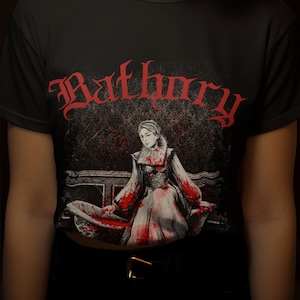 Op de afbeelding: Zwart T-shirt met een rode grafische print van een vrouw in een witte jurk met bloedspatten. De tekst "Bathory" is in rood boven de grafische print gedrukt.