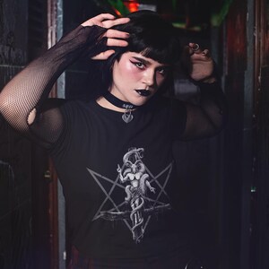 May include: A person wearing a black long-sleeved shirt with a pentagram and a creature design. The shirt has a mesh overlay on the sleeves. The person is wearing a black choker necklace with a silver pendant.