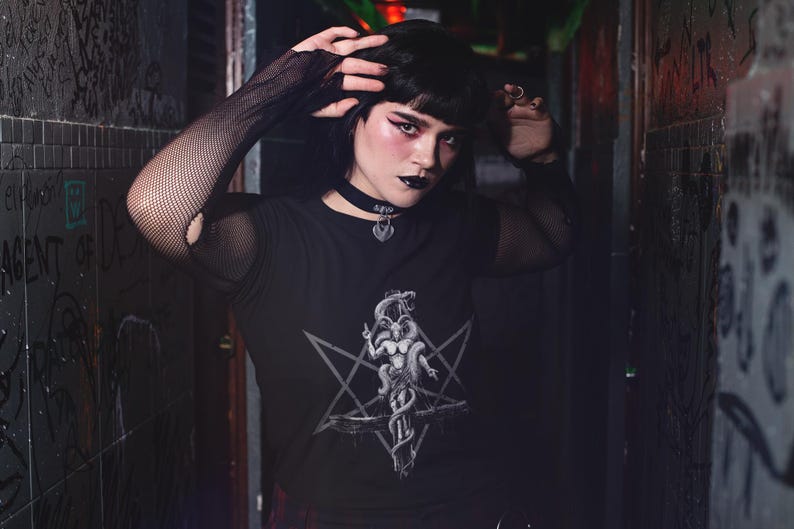 May include: A person wearing a black long-sleeved shirt with a pentagram and a creature design. The shirt has a mesh overlay on the sleeves. The person is wearing a black choker necklace with a silver pendant.