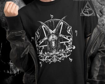 May include: Black t-shirt featuring a detailed white illustration of a goat's head within a pentagram, surrounded by ornate floral elements and symbols. The text "AMERICAN WITCH" and a triangular logo are visible on the upper right. The person is wearing a black leather jacket.