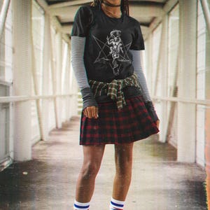 May include: A person wearing a black t-shirt with a white graphic print of a pentagram and a figure. They are also wearing a red and black plaid skirt, white and blue striped socks, and black combat boots.