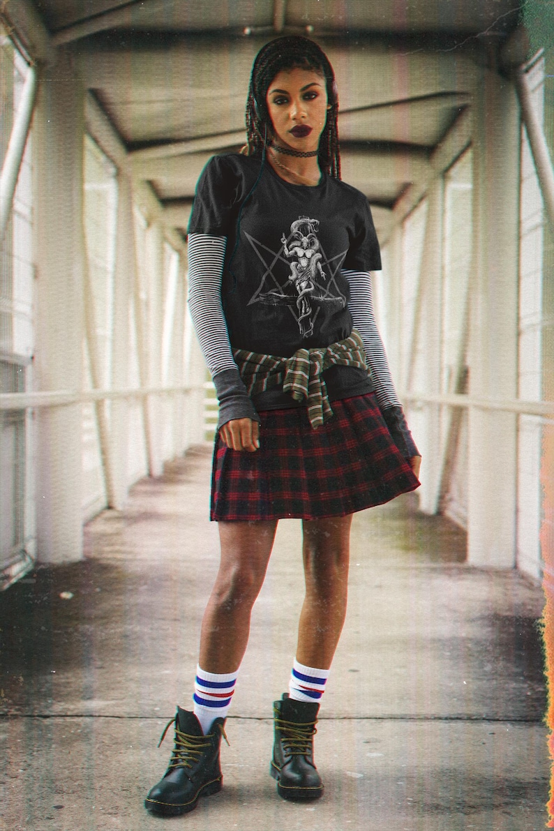May include: A person wearing a black t-shirt with a white graphic print of a pentagram and a figure. They are also wearing a red and black plaid skirt, white and blue striped socks, and black combat boots.