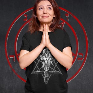 May include: A black t-shirt featuring a white graphic print of a goat-like figure with horns and a snake wrapped around it. The figure is standing on a cross with a pentagram behind it.