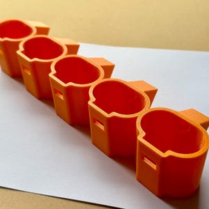 May include: Five orange plastic holders arranged diagonally. Each holder has a cylindrical interior and a rectangular exterior with a small rectangular opening. The holders are designed to hold a cylindrical object. The background is a light tan colour.