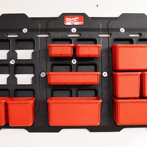 Può includere: Un sistema di stoccaggio nero a parete con contenitori rossi. Il sistema presenta un design a griglia con più scomparti e il logo "Milwaukee PACKOUT". I contenitori sono di varie dimensioni, offrendo opzioni di stoccaggio versatili.