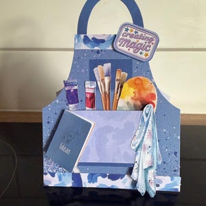 May include: A blue paper craft organiser shaped like a tote bag with a handle. It features a pocket containing paint tubes, brushes, and a palette. A small blue notebook labelled Ideas and a patterned cloth are attached. A patch reads creating Magic.
