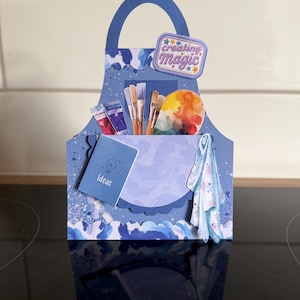 May include: A blue paper craft shaped like an apron with a handle, featuring a pocket containing paint tubes, brushes, and a palette. A small notebook labelled Ideas and a cloth are attached. The text creating magic is printed on a small sign.