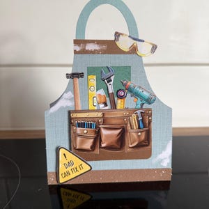 May include: A die-cut card shaped like a light blue apron featuring a brown leather tool belt graphic. The design includes illustrations of a hammer, wrench, drill, level, and safety glasses. A yellow triangular sign reads DAD CAN FIX IT.