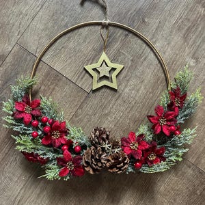 May include: A gold metal wreath adorned with red poinsettias, red berries, and pinecones. The wreath features green faux pine needles and a gold star pendant. A festive holiday decoration.