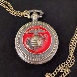 Op de afbeelding: Een vintage zakhorloge met een goudkleurige ketting. De wijzerplaat toont het embleem van het United States Marine Corps op een rode achtergrond, omgeven door een grijze rand. De ketting heeft een klassiek schakelontwerp.