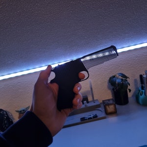Könnte beinhalten: Ein schwarzer Kamm in Form einer Pistole. Der Kamm hat einen Griff und eine Reihe von Zähnen. Der Kamm wird in einer Hand vor dem Hintergrund eines Schreibtisches und einer Wand gehalten.