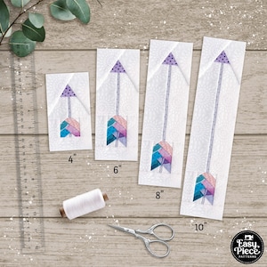 May include: Four different sizes of arrow quilt blocks with a purple, pink, blue, and teal geometric pattern. The blocks are 4 inches, 6 inches, 8 inches, and 10 inches wide. The blocks are on a white wooden surface with a ruler and a spool of white thread.