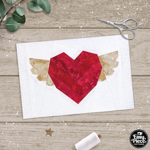 May include: A red heart with gold wings, made from fabric scraps, is displayed on a white background. The heart is a patchwork of red fabric squares, and the wings are made from gold fabric scraps. The heart is a symbol of love and hope.