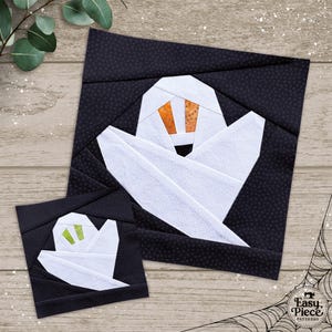 以下が含まれることがあります： 木目調の背景に、大小2つの布製ゴーストのデザイン。ゴーストは白で、オレンジ色または緑色の目があり、水玉模様の黒い背景に配置されています。デザインは正方形で、パッチワークプロジェクトの一部であるようです。