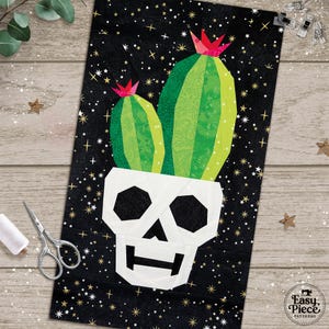 May include: A rectangular fabric quilt block featuring a white skull with black eye sockets and a mouth, topped with two green cacti with pink flowers. The background is black with a pattern of small white stars. The text Easy Piece Patterns is visible.