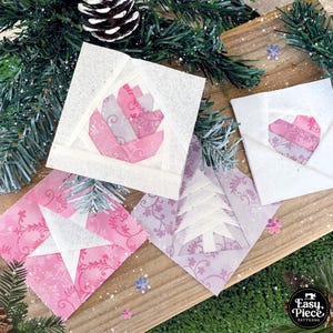 Può includere: Quattro ornamenti natalizi in carta a pezzi rosa e bianchi, tra cui una stella, un albero, un cuore e un fiore. Gli ornamenti sono su una superficie di legno con uno sfondo verde.