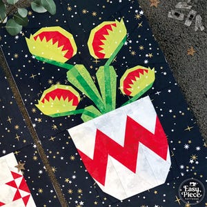 May include: A fabric quilt block featuring a dark navy background with gold stars. The design depicts a potted plant with three green and red Venus flytrap heads. The pot is white with a red zigzag pattern. A logo reads Easy Piece Patterns.