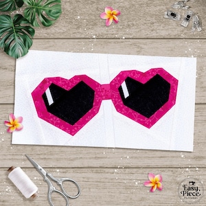May include: A quilt block pattern featuring a pair of heart-shaped sunglasses with bright pink frames decorated with a white floral vine print and solid black lenses with white rectangular highlights, set against a light gray patterned background.