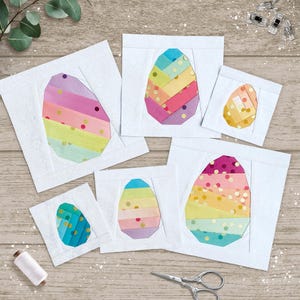 Könnte beinhalten: Sechs Stoff-Osterei-Designs in verschiedenen Größen. Jedes Ei hat ein Patchwork-Design mit bunten horizontalen Streifen und goldenen Punkten, vor einem weißen quadratischen Hintergrund. Eine Garnrolle und eine Schere sind ebenfalls sichtbar.