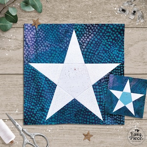 May include: A large square fabric block with a white star design on a blue background, alongside a smaller version. The star is made of white fabric. The background has a speckled pattern. Sewing tools and a spool of thread are visible.