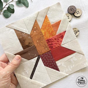 Op de afbeelding: Een vierkant stoffen quiltblok met een centraal esdoornbladontwerp, samengesteld uit verschillende herfstkleurige stoffen met patronen in bruin-, oranje- en roodtinten, tegen een crèmekleurige achtergrond met drie houten knopen.