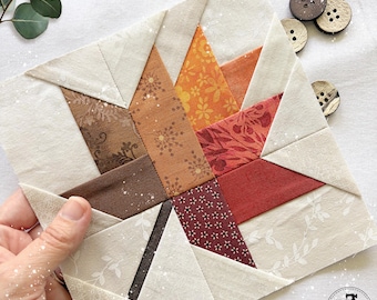 May include: A square fabric quilt block featuring a central maple leaf design pieced together from various autumn-colored patterned fabrics in shades of brown, orange, and red, set against a cream-colored background with three wooden buttons.