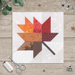 Puede incluir: Un bloque de edredón cuadrado con un diseño de hoja estilizada en tonos naranja, rojo y marrón, sobre un fondo blanco con un patrón floral. La hoja está compuesta por varias telas estampadas. Se ven accesorios de costura, como tijeras e hilo.