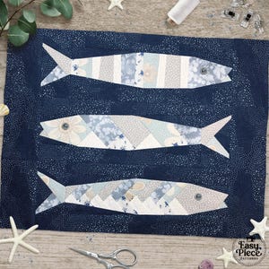 May include: A navy blue fabric with three fish appliqués. The fish are pieced together with various blue, white, and gray patterned fabrics. The fish have a floral eye detail. The background fabric has a small white dot pattern. The image includes sewing notions.