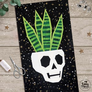 May include: A rectangular fabric panel featuring a white skull-shaped planter containing four green striped leaves. The background is black with gold star accents. The text "Easy Piece Patterns" is printed in the bottom right corner.