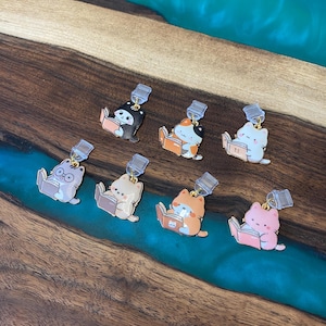 May include: Eight colorful cat bookmarks with different colored books and cats. The cats are reading books and have different expressions on their faces. The bookmarks are made of metal and have a clear plastic clip on the top.