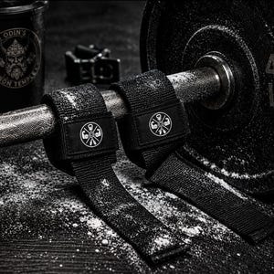 May include: A pair of black weightlifting straps featuring a circular logo with icons of a kettlebell, a barbell, and flexed biceps. The straps are wrapped around a metal barbell next to a 45 LB weight plate and a shaker bottle labelled Odin's Iron Temple.