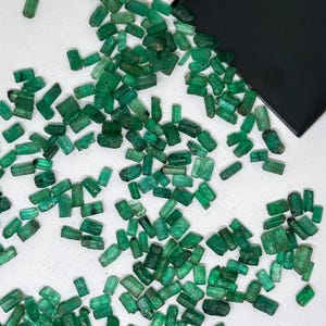 Op de afbeelding: Een grote hoeveelheid kleine, ruwe, groene smaragd edelstenen. De smaragden zijn verspreid over een wit oppervlak.