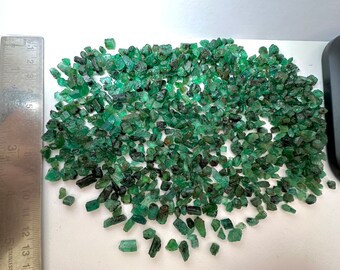 May include: A large pile of small, green emerald crystals. The crystals are various shapes and sizes, with some being long and thin, while others are more rounded. The crystals are scattered on a white surface, with a ruler in the background for scale.