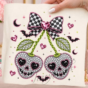 May include: A cream-colored fabric featuring a graphic of two skull-shaped cherries with heart-shaped eyes and stitched mouths. The cherries are topped with green stems and a large black and white checkered bow with a pink heart center, surrounded by small pink hearts, black crescent moons, and black bats.