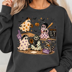 May include: A dark grey crewneck sweatshirt featuring a graphic print of ghosts and raccoons playing baseball on a field. The design includes a scoreboard reading SPOOKY FIELD, a pumpkin, bats, and text that reads #1, BOO!, and GO GHOSTS!.